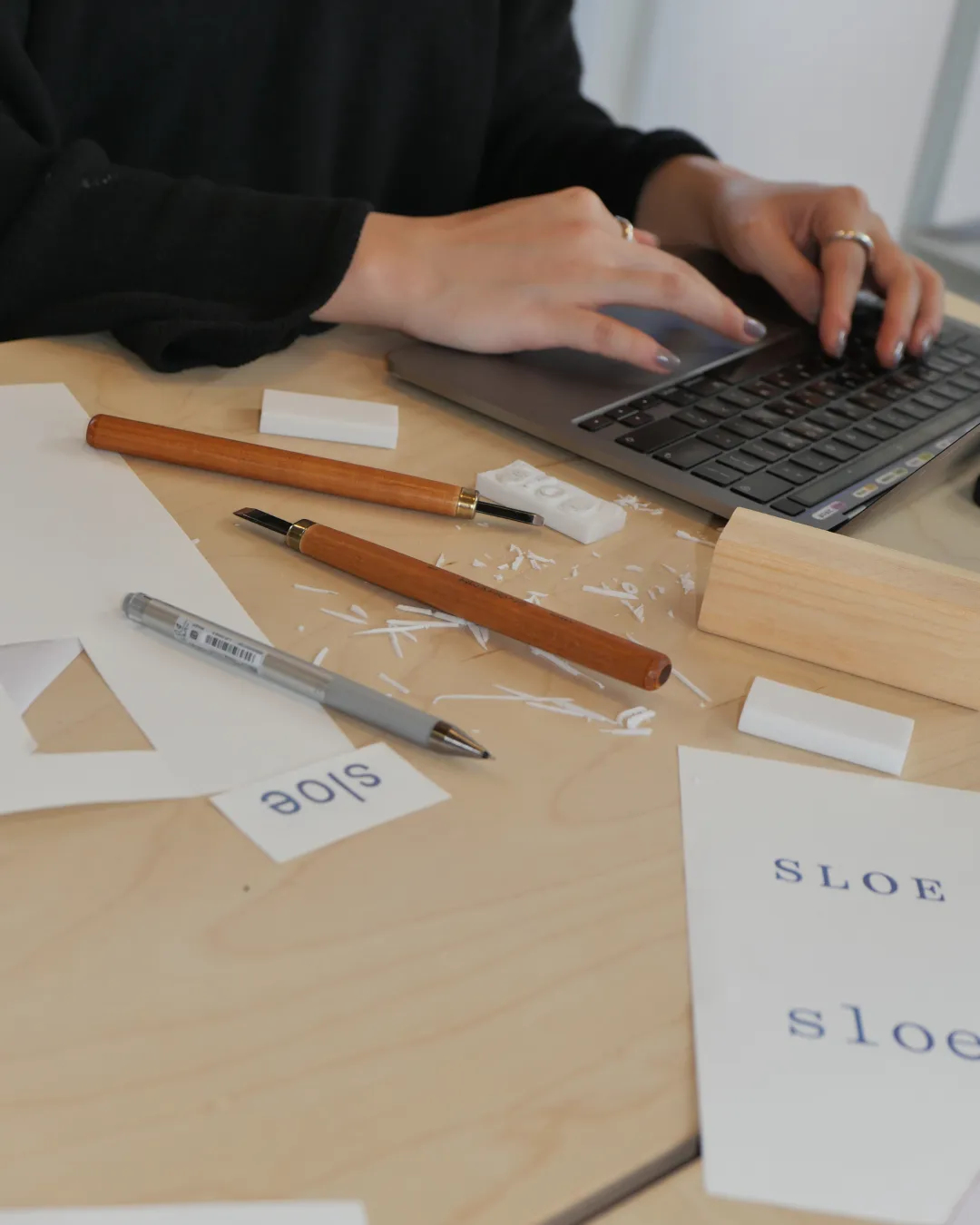 Hands typing on a laptop beside pens and printed type samples